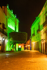 A street with a green tree in the middle and buildings on both sides. The street is lit up at night. Historic center of Queretaro, colonial architecture, decorations for the celebration of Mexico's In