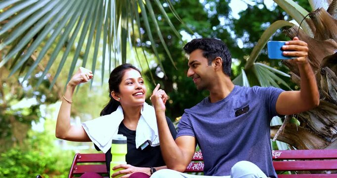 Indian young couple taking selfie after exercising in park sitting on bench enjoying post workout relaxation, capturing happy fitness moment together with smartphone in morning sunlight in garden