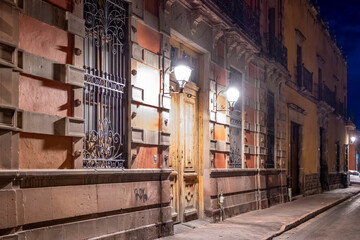 A street at night with a yellow door and a light on the front. The street is empty and quiet....