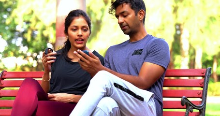 Indian beautiful young couple checking fitness app on smartphone sitting on bench in park outdoor during sunny day, tracking health progress, wellness goals, and sharing workout motivation - Powered by Adobe