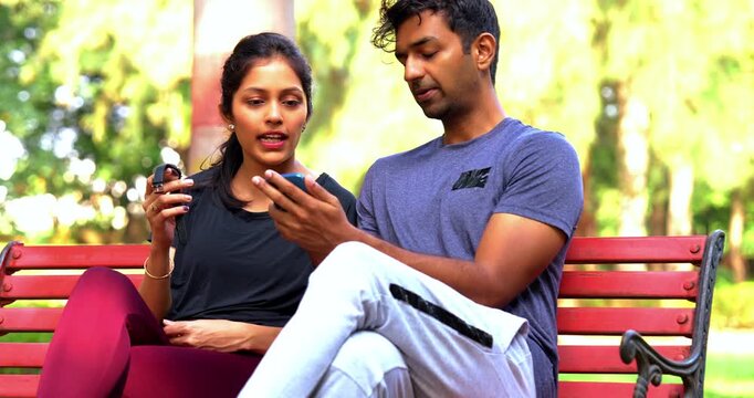Indian beautiful young couple checking fitness app on smartphone sitting on bench in park outdoor during sunny day, tracking health progress, wellness goals, and sharing workout motivation
