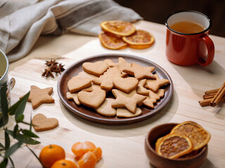 Wooden platter with baked gingerbread shapes is surrounded by seasonal spices, oranges, and a mug...