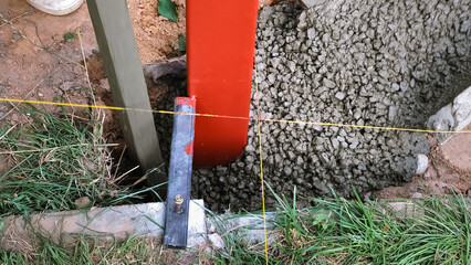 Naklejka premium A builder pours a wet concrete from a mixer truck into a foundation. Pouring cement into pit with pillar. Foundational work. Workers control a mix flows through the gutter. Building industry.