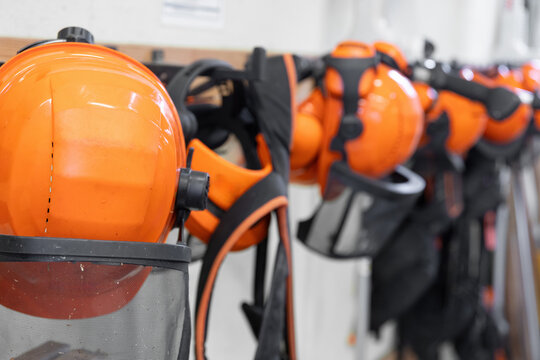 Reihe von orangefarbenen Schutzhelmen mit Visier und Geh&ouml;rschutz h&auml;ngt ordentlich an einer Wand. Das Motiv steht f&uuml;r Arbeitssicherheit, Forst- und Handwerksberufe sowie professionelle Schutzausr&uuml;stung