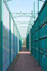 A narrow, open-air corridor formed by green metal structures and fences made of mesh and boards, with a perspective stretching into the distance.