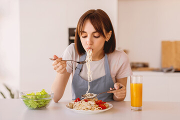Young housewife tasting spaghetti and vegetable salad, enjoying delicious lunch at home, sitting at table in modern kitchen interior, free space