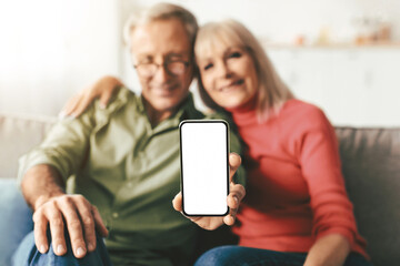 Elderly Couple Showing Phone Empty Screen To Camera Advertising Mobile Application For Your Smartphone Sitting On Sofa At Home. Selective Focus On Cellphone Display. Great App Concept. Mockup
