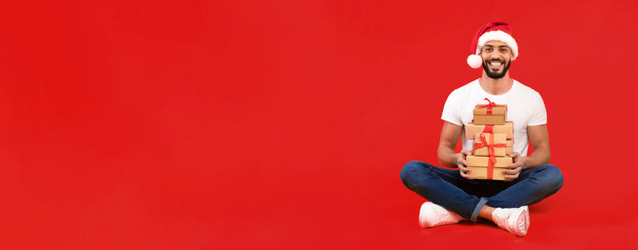 Joyful Arabic Family Posing With Christmas Gifts. Happy Man Wearing Santa Hat Holding Wrapped Presents Boxes Sitting With Wife And Daughter Over Yellow Background. Studio Shot
