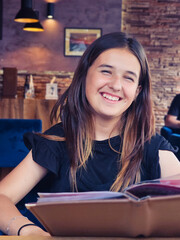 Smiling teenage girl sitting in a cozy cafe, holding a menu and enjoying a relaxed moment indoors.
