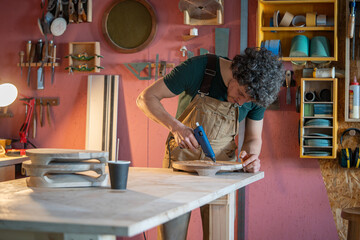 Woodworker apply melted silicone along edges of imperfections in crafted timber before resin repair. Crack stabilization, wood surface defect correction, furniture flaw preventive wood restore