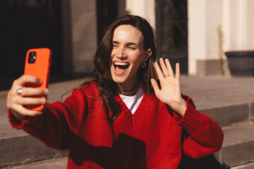 Beautiful smiling long brunette hair woman taking selfie and waving hand hi, hello, bye while sitting on old museum stairs. Woman wear red sweater. Student or businesswoman.