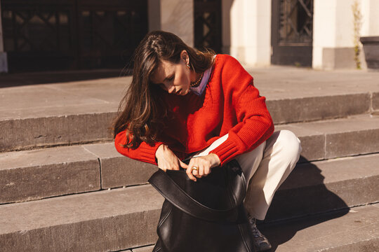 Outdoor portrait of young attractive woman with brunette hair take out a notebook from her backpack, serious and have new ideas for note. Sitting on stairs, wears red sweater, black leather bag. - Powered by Adobe