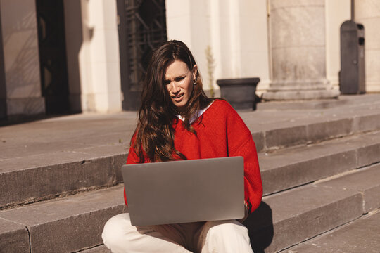 Angry shocked young woman looking at laptop screen, sitting on steps outdoors, she reading bad news in email or message, financial problem, wear red sweater.