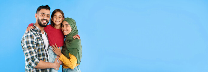 Muslim Parents Embracing Little Daughter Smiling To Camera Posing Together Standing Over Blue...