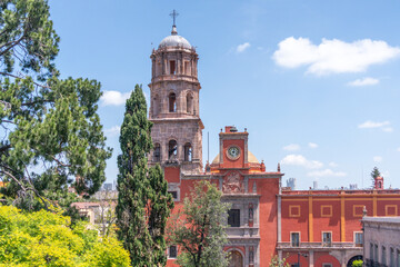 A large red building with a clock tower and a spire. The building is surrounded by trees. Historic center of Queretaro, colonial architecture, decorations for the celebration of Mexico's Independence 