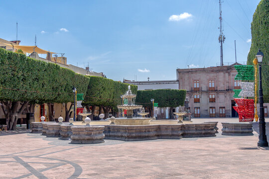 A courtyard with a fountain and trees. The fountain is surrounded by stone pillars. Historic center of Queretaro, colonial architecture, decorations for the celebration of Mexico's Independence Day.
