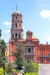 A large red building with a clock tower and a spire. The building is surrounded by trees. Historic center of Queretaro, colonial architecture, decorations for the celebration of Mexico's Independence 