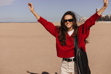 Happy brunette long hair woman dancing on the beach coast walking on spring or autumn sunny day. Woman wear red sweater, black sunglasses and bag on shoulders. Happy motion girl, hands side up.
