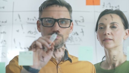 Professional team members brainstorming strategy, writing notes on transparent whiteboard with colorful sticky papers during collaborative meeting. Business people concept