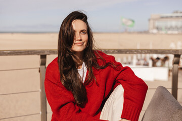 Portrait of charming brunette long hair woman wear red sweater. Girl sits on stone chair on coast and enjoys spring or autumn day. Calm zen woman look at coast. © zvkate