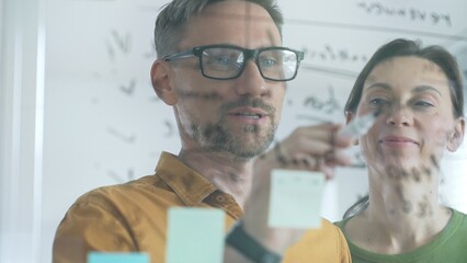 Professional team members brainstorming strategy, writing notes on transparent whiteboard with colorful sticky papers during collaborative meeting in office. Business people at work