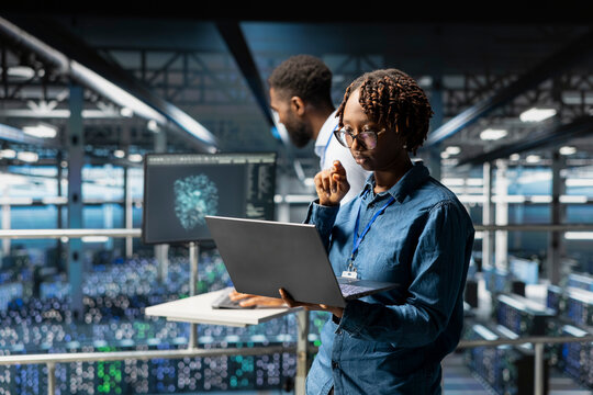 African american female expert doing maintenance tasks on laptop in data center, ensuring optimal performance. Engineer reviewing diagnostics and applying patches to server hub gear. - Powered by Adobe