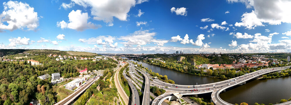 Wide aerial panorama of highway interchange with multiple roads, river and green hills under blue sky, modern transport infrastructure and mobility network in scenic landscape.