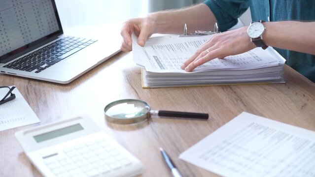 Close-up of man hands reviewing financial documents with magnifying glass. Business work process in bookkeeping office. Taxes and audit - Powered by Adobe