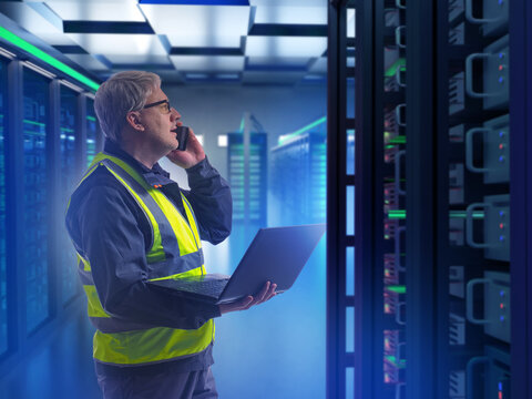 Engineer in safety vest with laptop and phone in a server room, symbolizing IT, data management, and cybersecurity operations. Information Technology, Data.