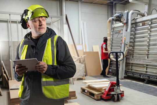 Supervisor in safety vest and helmet with tablet controlling woodworking production in factory, worker operating cutting machine. Manufacturing, warehouse