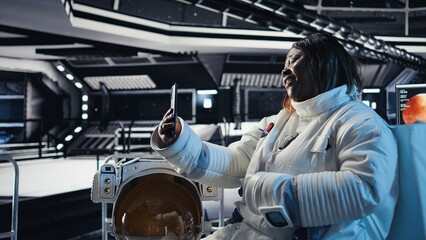 Happy female cosmonaut doing videocall with family using smartphone during galaxy expedition. Africna american woman doing remote call with loved ones using mobile phone in spaceship