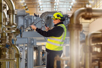 Engineer in helmet and vest using measuring device to check industrial machinery in factory,...