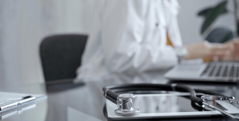 Close-up of doctor's desk with stethoscope and tablet computer. Physician is using a laptop at the background. Medicine concept