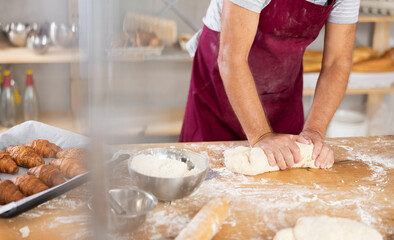 Close-up of professional male baker hands in dark red apron kneading dough on flour-dusted wooden table in artisan bakery, highlighting craftsmanship of traditional breadmaking