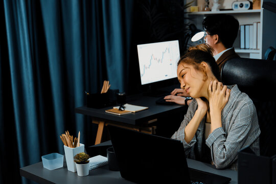Businesswoman working on desk with stretching arm up and down manner with body health ache of strain overwork while coworker trading stock market on website on pc at late over night time. Infobahn. - Powered by Adobe