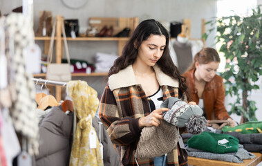 Armenian woman stands in a warm jacket and carefully chooses a hat for autumn and winter. Buyer looks at knit accessories against background of a woman who chooses sweaters at the counter of a store