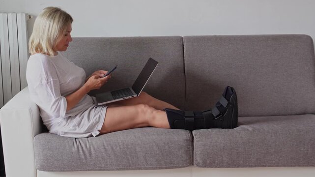 Woman with fractured leg sitting on the sofa with orthopedic medical Walker boot, support splint foot, speaking on smartphones and working on the laptop. Remote jobs during the recovery from the