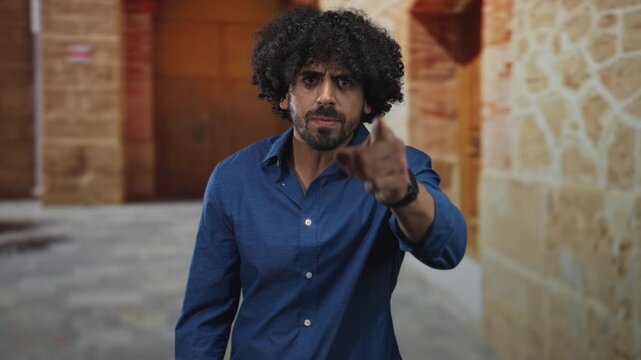 Man in blue shirt pointing finger forward with visible hand and stern expression in old town street; anger accusation.