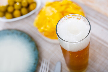 Glass of chilled foam frothy beer with snacks - portion of yellow crispy potato chips on small plate and bowl with delicate green olives