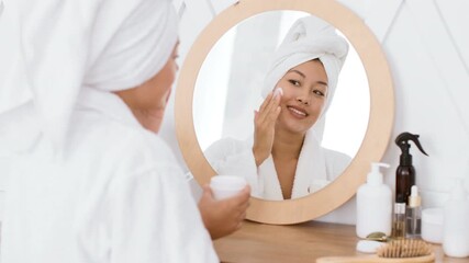 In a serene home setting, a young Asian woman pampering herself applies skincare products while smiling in the mirror, embracing a soothing beauty ritual.