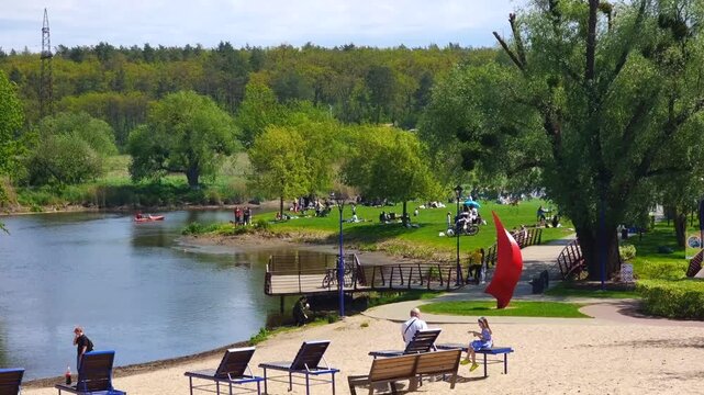 People enjoying a beautiful sunny day in a city park, resting on benches and lounge chairs on a sandy beach by the river, with lush green trees and a forest in the background