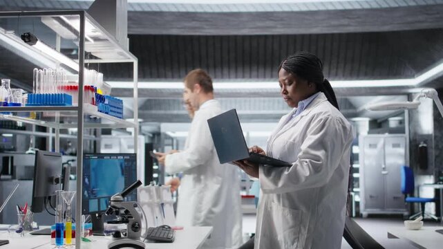 Black female researcher in a clinical research lab studies experiment data with a laptop. Showcasing biotechnology, genetics research and pharmacology for progress in healthcare. Camera B.