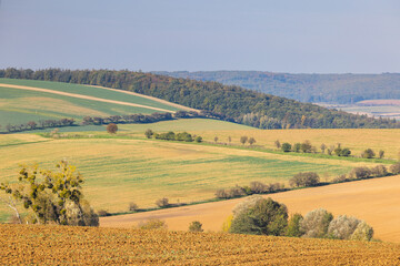 Rolling hills and farmland under a clear sky create a peaceful rural landscape. Textured fields in warm earth tones are visible. Moravia region of Czech Republic, Europe.
