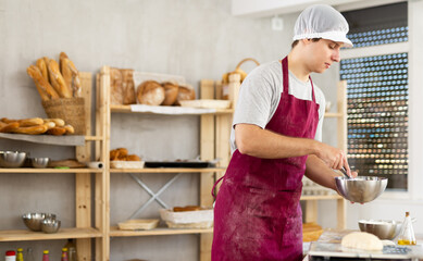 Young male baker in uniform whipping dough in bowl