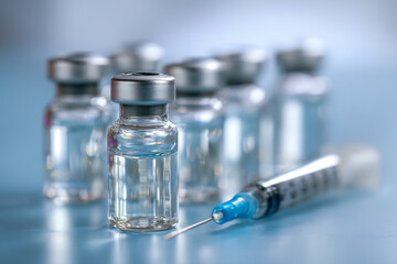 Medical vials containing clear liquid with hypodermic syringe placed on reflective surface in a clinical setting for vaccination or injection purposes