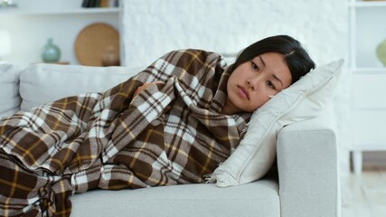 A young Asian woman is wrapped in a cozy blanket, resting comfortably on a sofa, got sick, suffering from fever. - Powered by Adobe