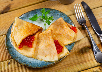 On plate are several rolled delicate pancakes with ruddy edge and spoonful red salmon caviar. Hearty snack is complemented by cup of fragrant coffee, tea