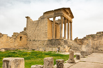 Fototapeta premium Well-preserved remnants of Capitol, historic temple with towering columns among ruins of ancient Roman settlement of Dougga in northern Tunisia on cloudy spring day..