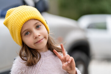 Young girl wearing yellow cap making a peace sign while smiling at camera. She looks calm and cheerful near blurred car in parking area during daytime