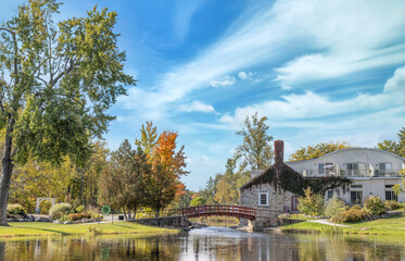 An old stone mill building and addition next to a small river, footbridge, trees and garden with fall colours, water in foreground, daytime, nobody